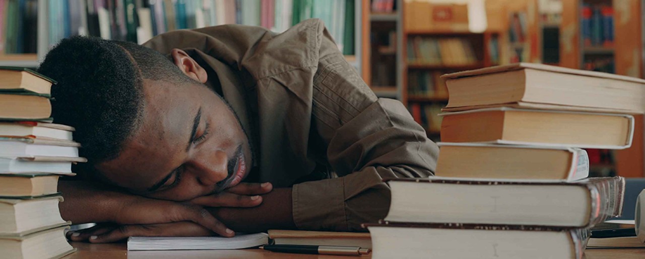 Black man resting his head on a desk full of books